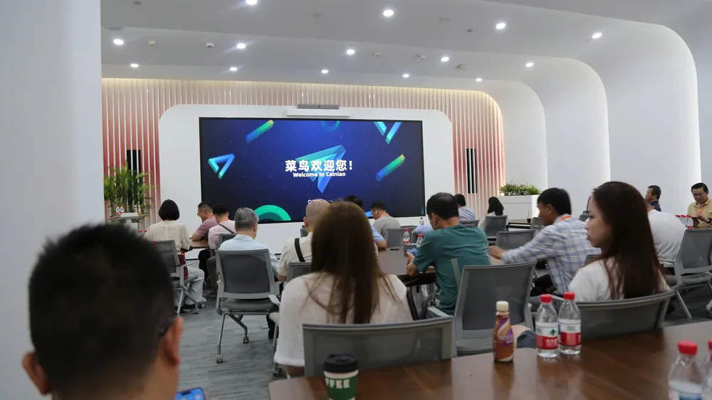 Newing delegation seated in the Cainiao hall; the large screen reads “菜鸟欢迎您！ Welcome to Cainiao”