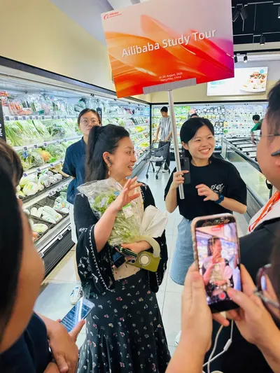 Delegation members holding an Alibaba Study Tour sign in the Hema supermarket area, one holding flowers and one filming on a phone